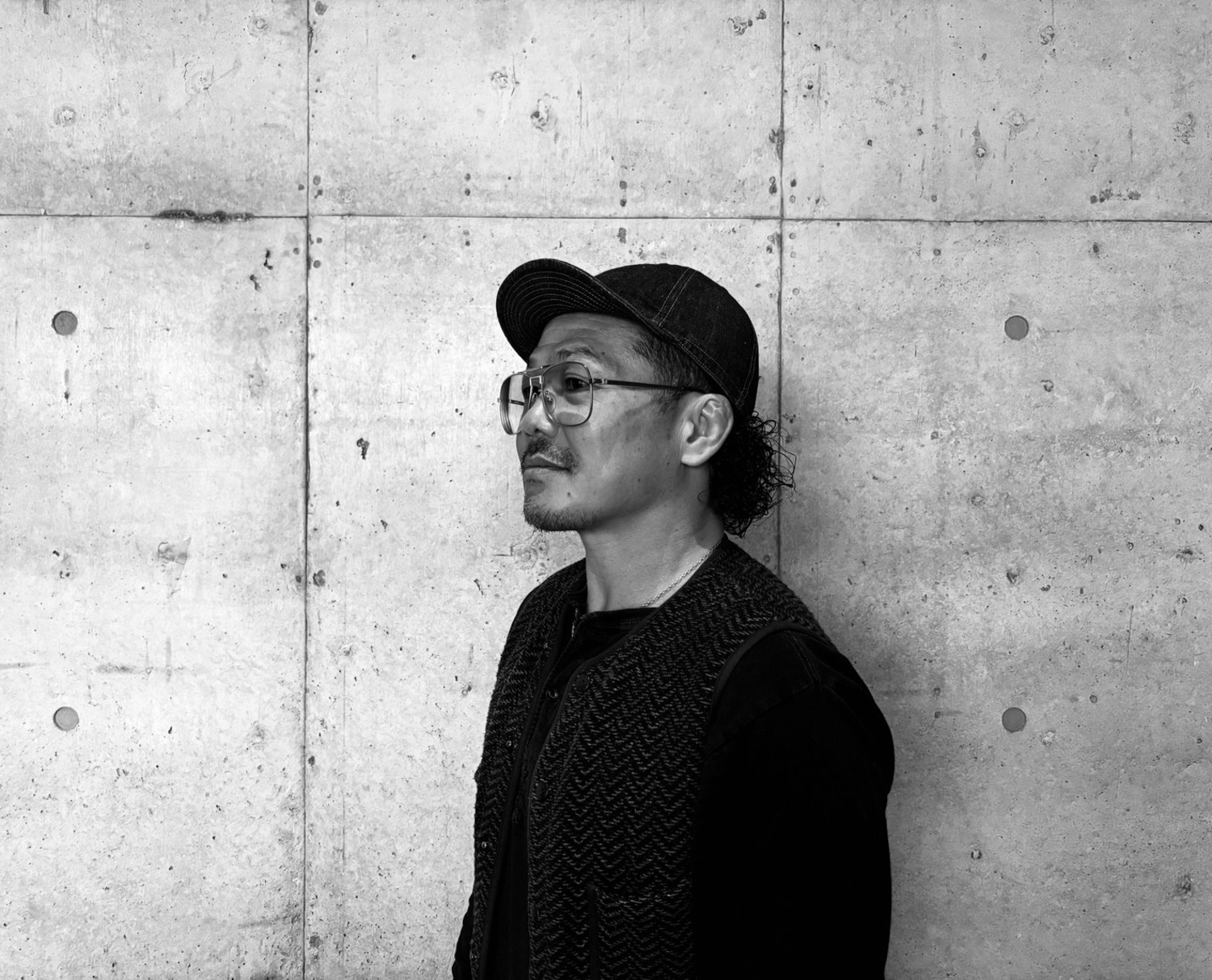 コンクリート壁の前に立つ橋本淳平さんの横顔ポートレート。キャップと眼鏡を着用したモノクロ写真