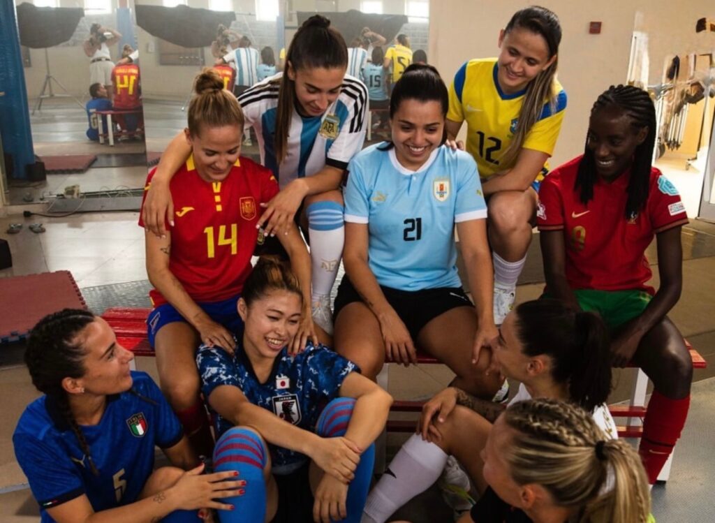 各国の代表ユニフォームを着た女子フットサル選手たちが室内で集まり、笑顔で会話する様子