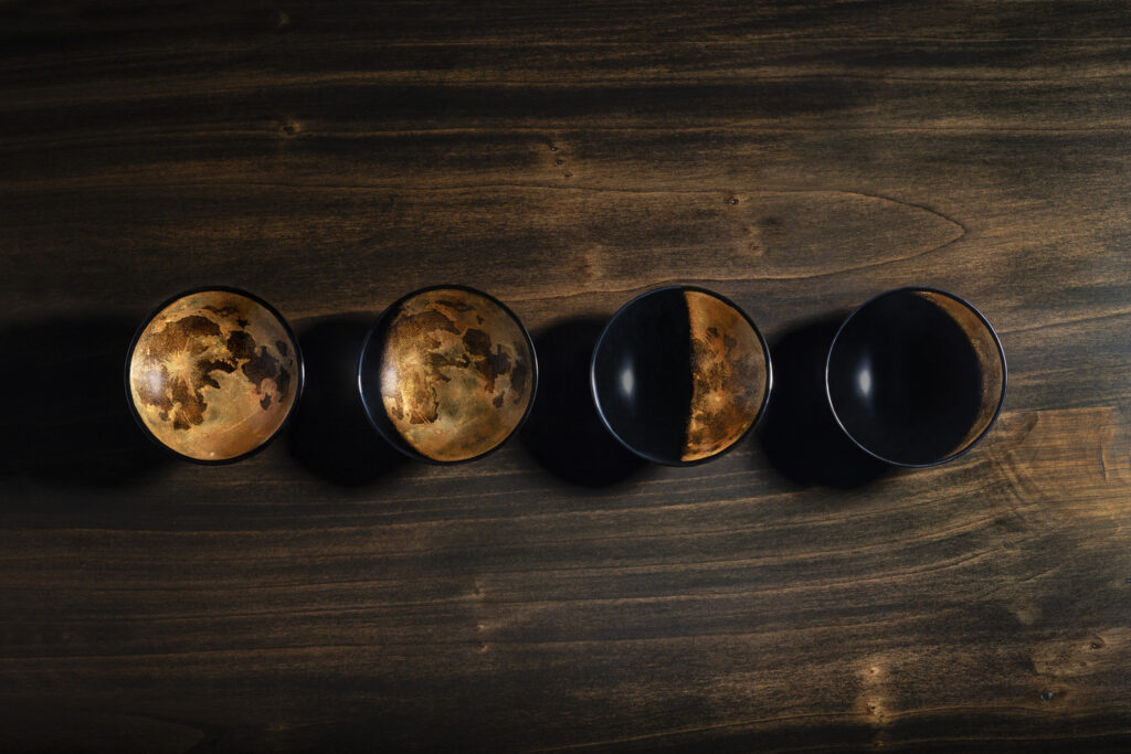 Maki-e Sake Cups “Lunar Calendar,” four black urushi sake cups with gold maki-e details arranged like phases of the moon on a dark wooden surface.