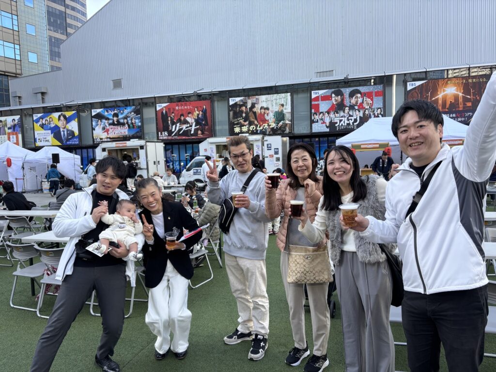 屋外イベント会場で、飲み物を手にした横尾俊成さんと参加者たちが笑顔で集まっている様子。