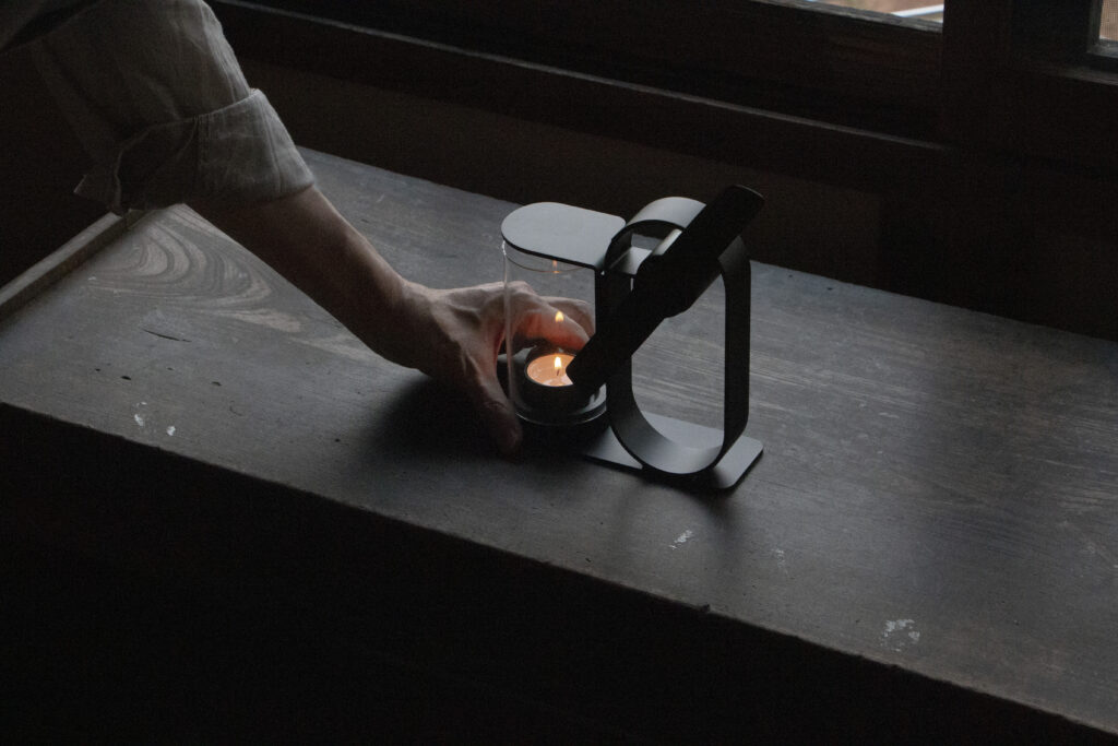 A hand resting against the base of a Lei non electric aroma diffuser with a lit candle on a wooden table.