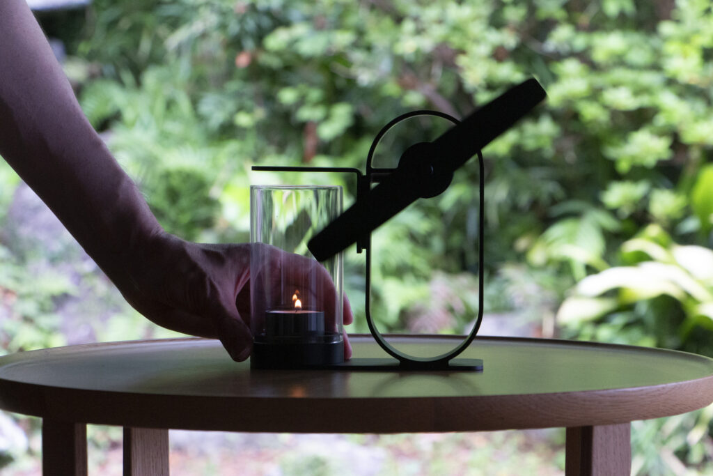A hand beside the Lei non electric aroma diffuser, with a lit candle beneath its propeller
