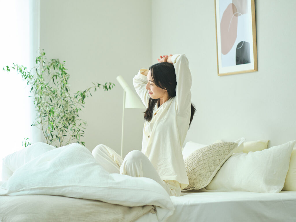 白い寝具に囲まれたベッドの上で、パジャマ姿の女性が目を閉じて伸びをしている朝の寝室。