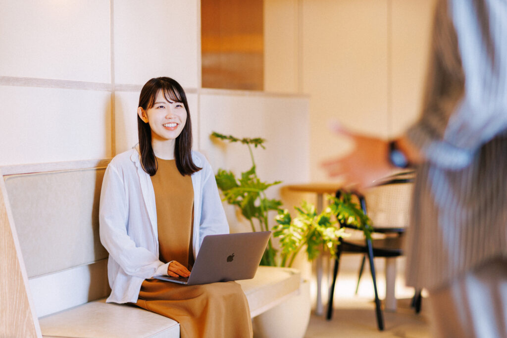 木と白を基調としたオフィスのラウンジで、ベンチに座りノートパソコンを膝に置いた女性が、同僚と笑顔で話している。
