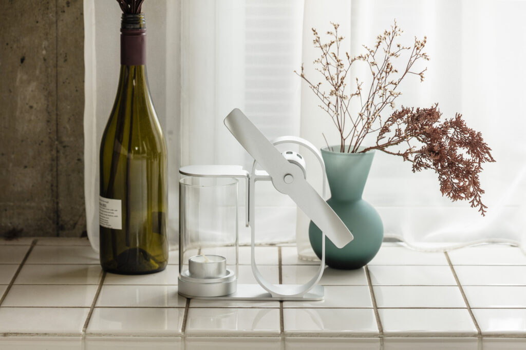 The Silver Lei non electric aroma diffuser on a white tiled counter by a sheer curtain, between a green glass bottle and a frosted vase of dried flowers.