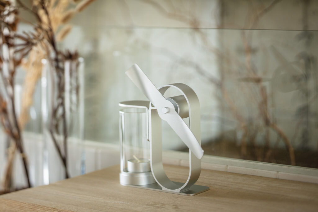 The Silver Lei non electric aroma diffuser on a wooden shelf beside a glass vase of dried branches, with the branches reflected in a glass panel behind it.
