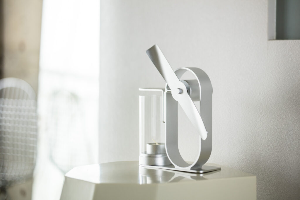窓から光が差し込む白い部屋で、八角形のサイドテーブルに置かれたSilverのLei non electric aroma diffuser。ガラスの筒の中にロウソクが入っている。
