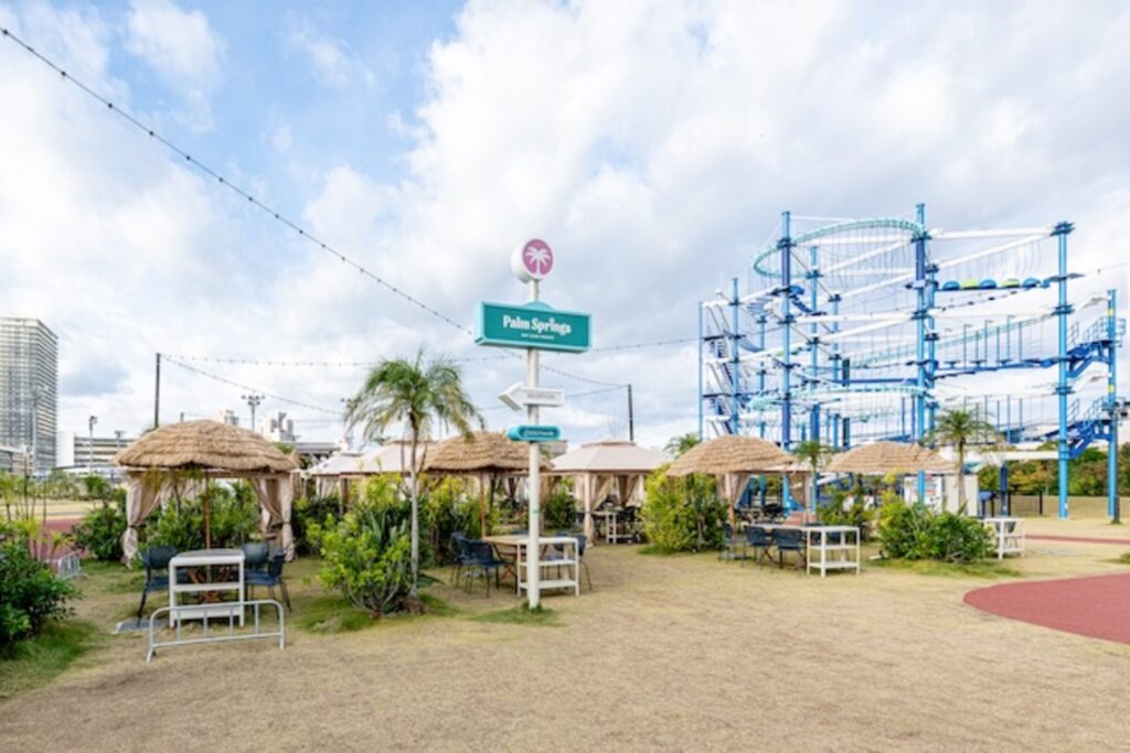 青空の下、サッチ屋根のパラソルとヤシの木が並ぶPalm Springsのパラソルサイト。奥にアスレチックのタワーが見える