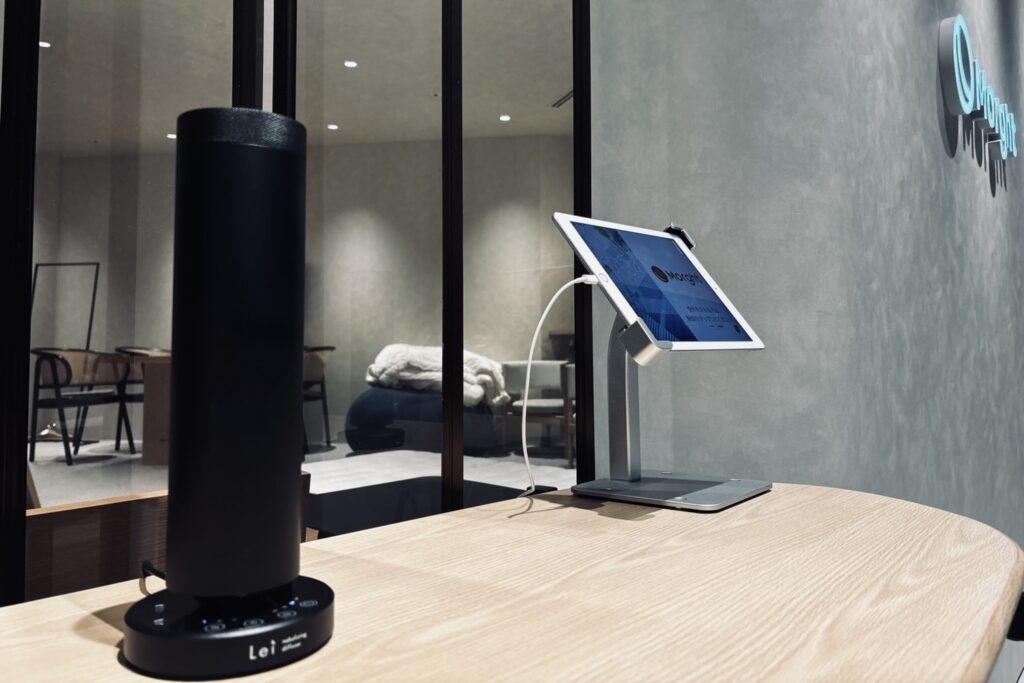株式会社Morghtのオフィス入口。木のカウンターに置かれた黒いLei業務用ディフューザーと受付用のタブレット。奥のガラス越しにベッドのある部屋が見える。