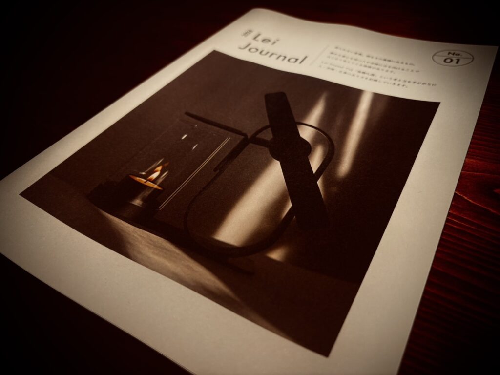 木のテーブルに置かれたLei Journal 紙面版 No.01の表紙。キャンドルの灯りとLei Non-Electric Aroma Diffuserの陰影を写した写真