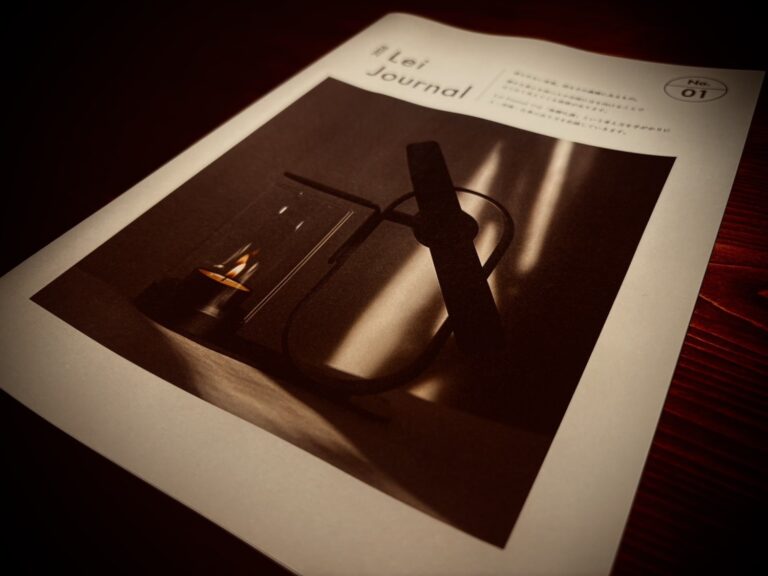 木のテーブルに置かれたLei Journal 紙面版 No.01の表紙。キャンドルの灯りとLei Non-Electric Aroma Diffuserの陰影を写した写真